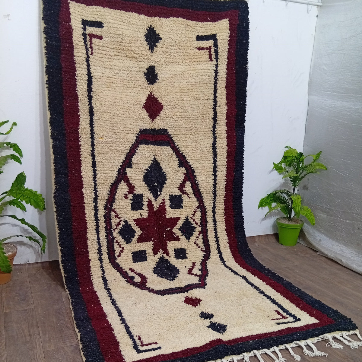 A traditional Moroccan-style wool rug with a cream base, dark navy blue borders, and a central red and navy geometric medallion, shown draped over a white wall on a wooden floor with potted plants.